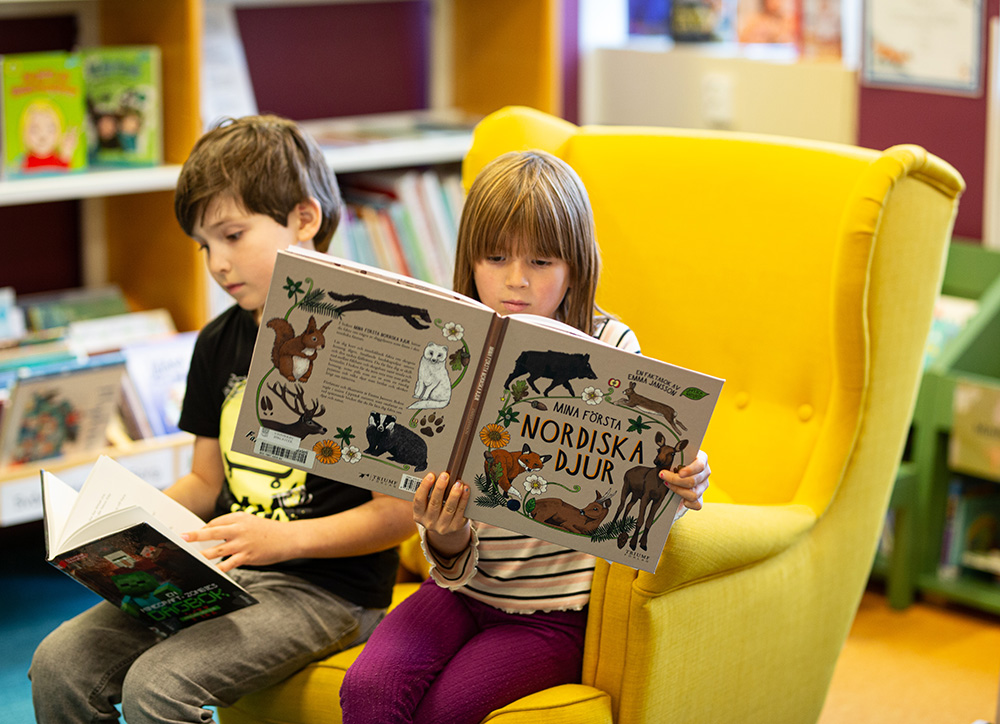 Två barn sitter tillsammans i en gul fåtölj på biblioteket och läser varsin bok